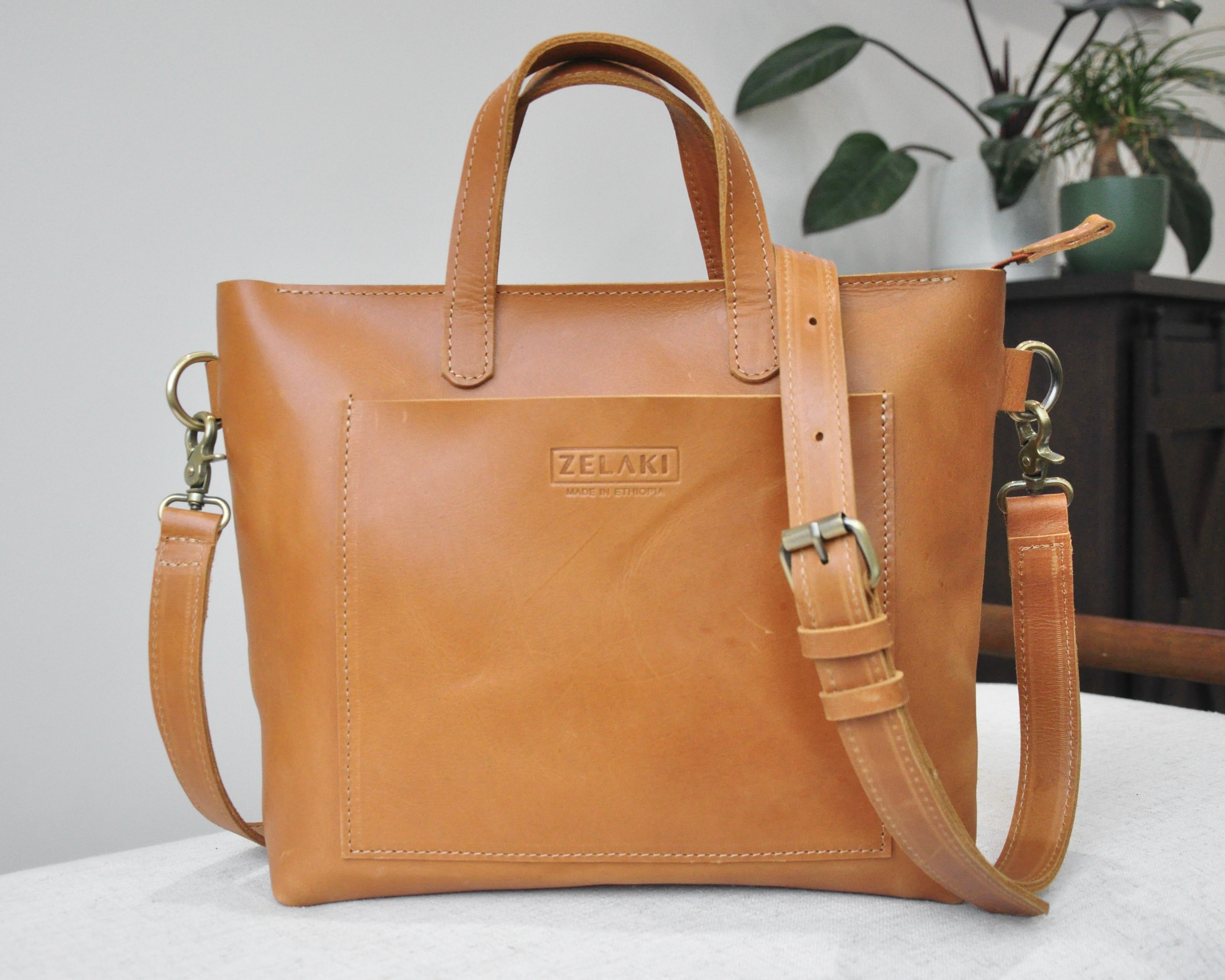 A medium sized, tan leather tote bag with a sleek, minimalist design featuring a top handle and adjustable crossbody strap