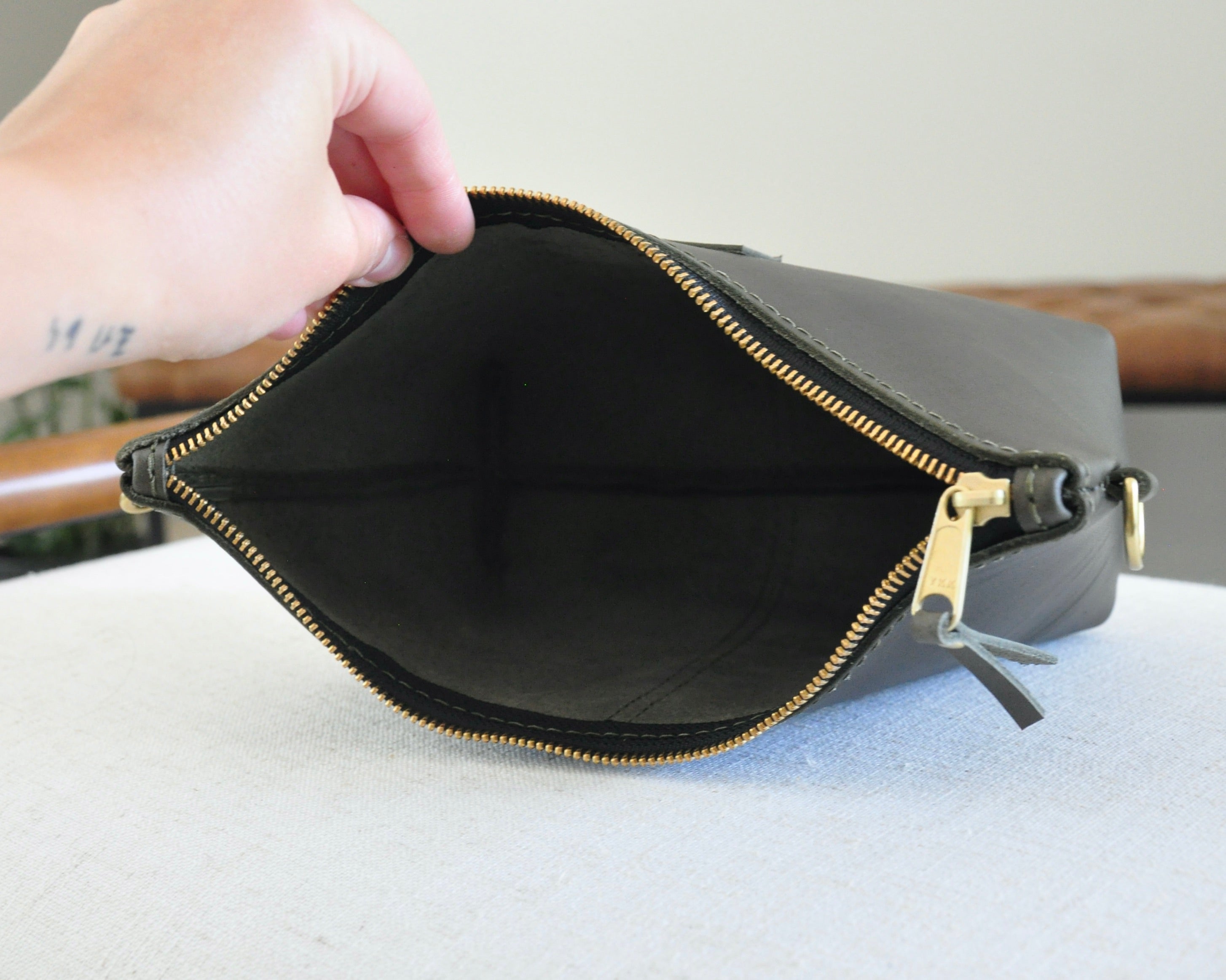 Hand holding a black clutch with gold zipper on a light gray surface