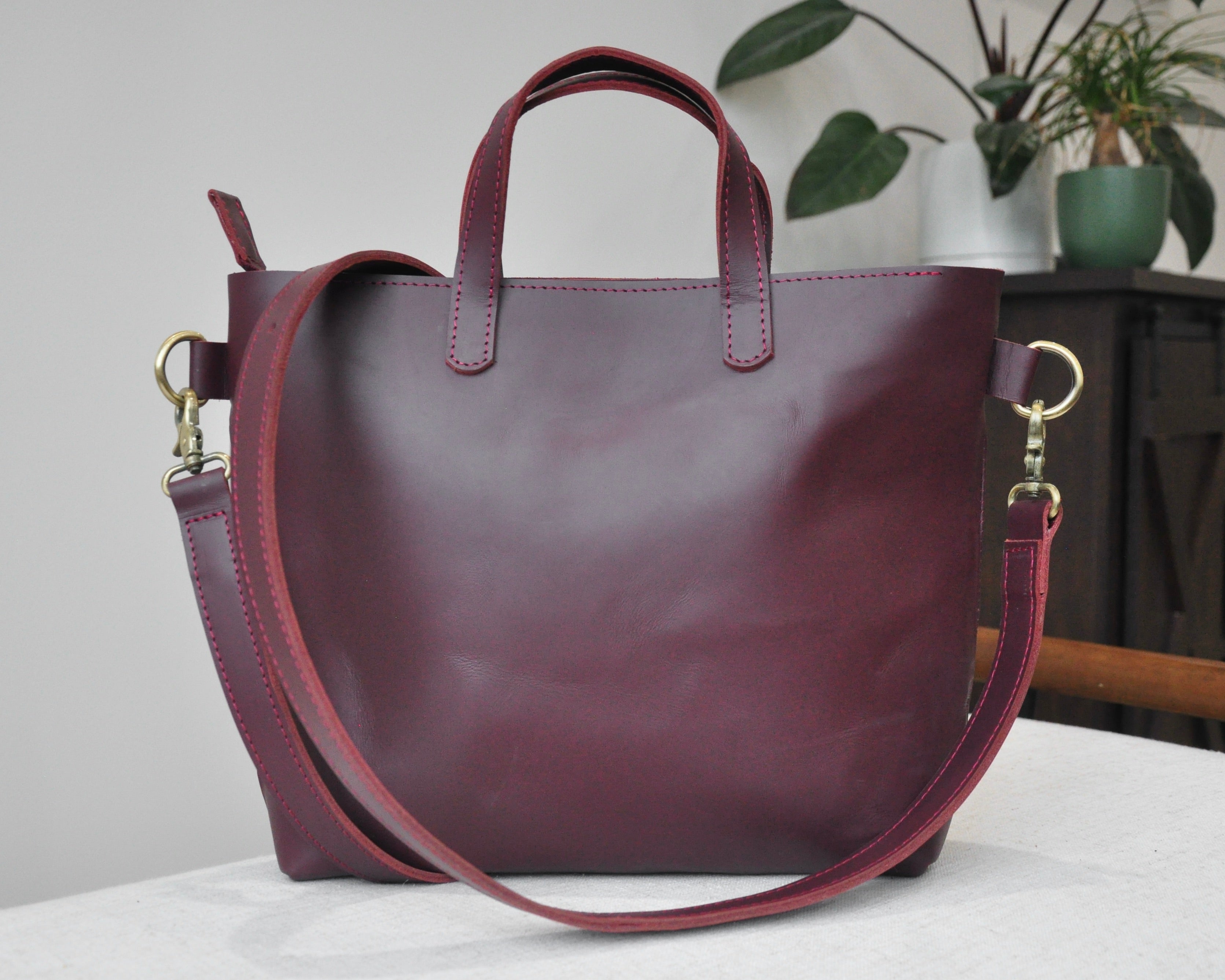 A medium sized, maroon leather tote bag with a sleek, minimalist design featuring a top handle and adjustable crossbody strap