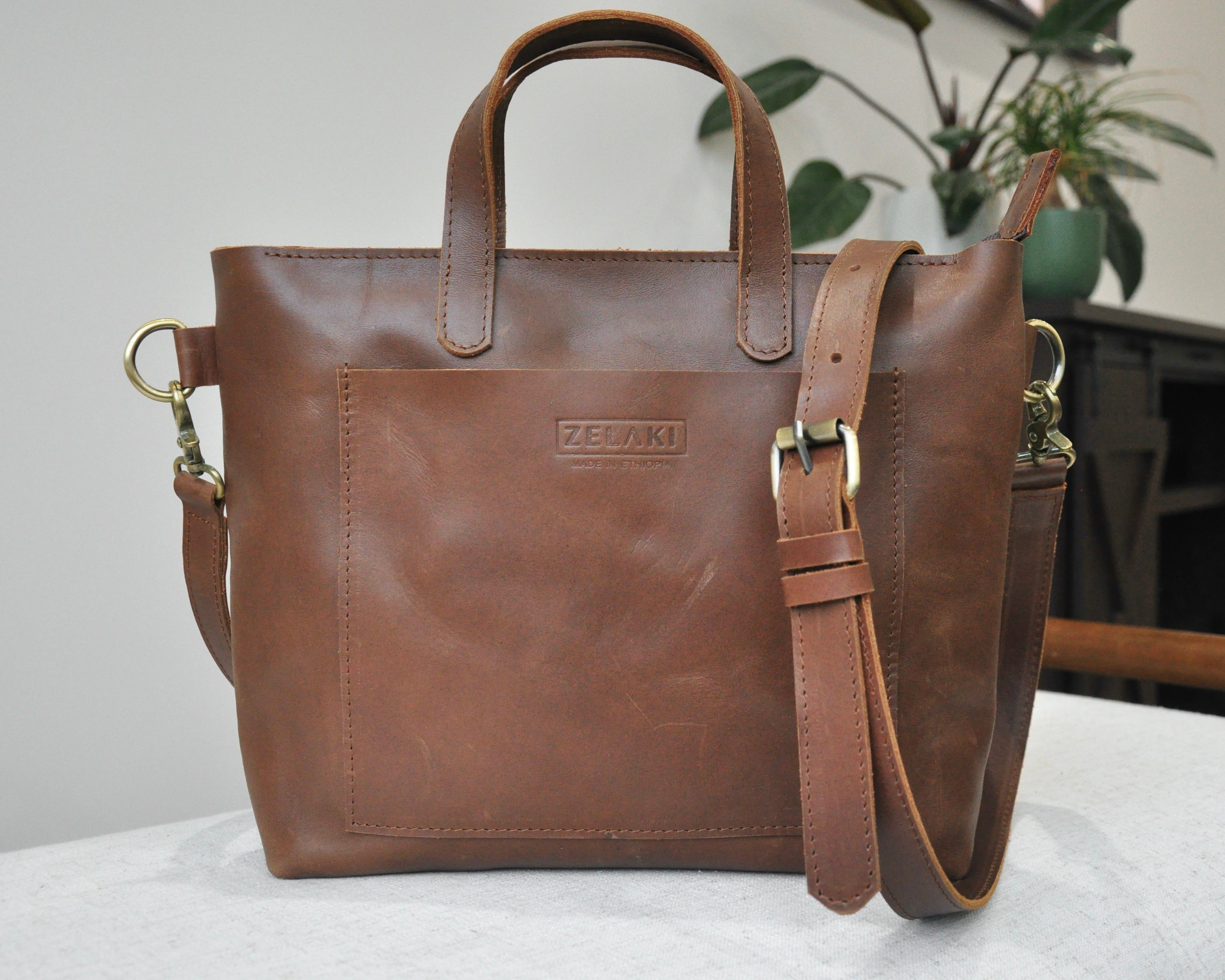 A medium sized, brown leather tote bag with a sleek, minimalist design featuring a top handle and adjustable crossbody strap