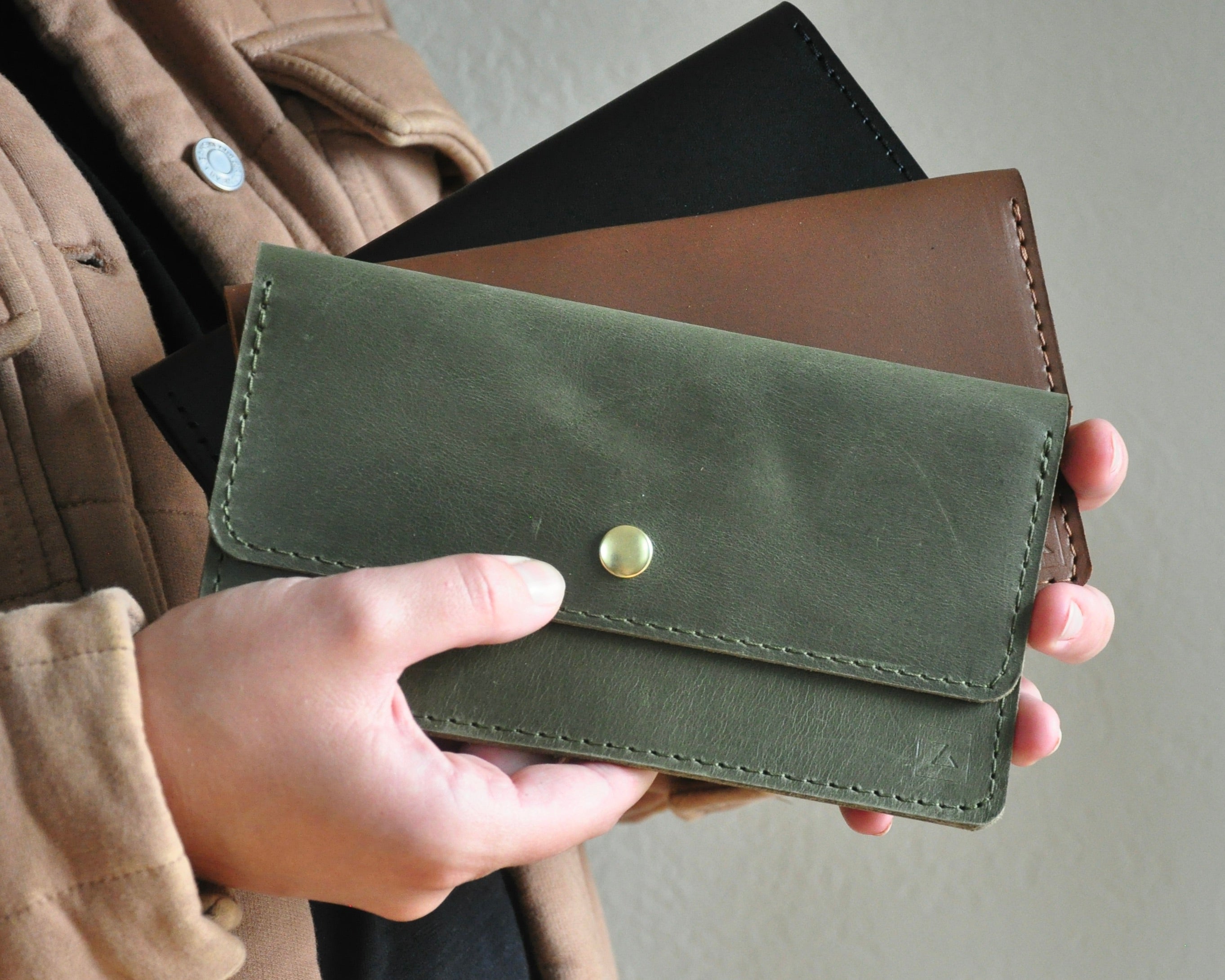 A person holding two wallet clutches in brown and green colors made of leather with a solid brass snap closure.