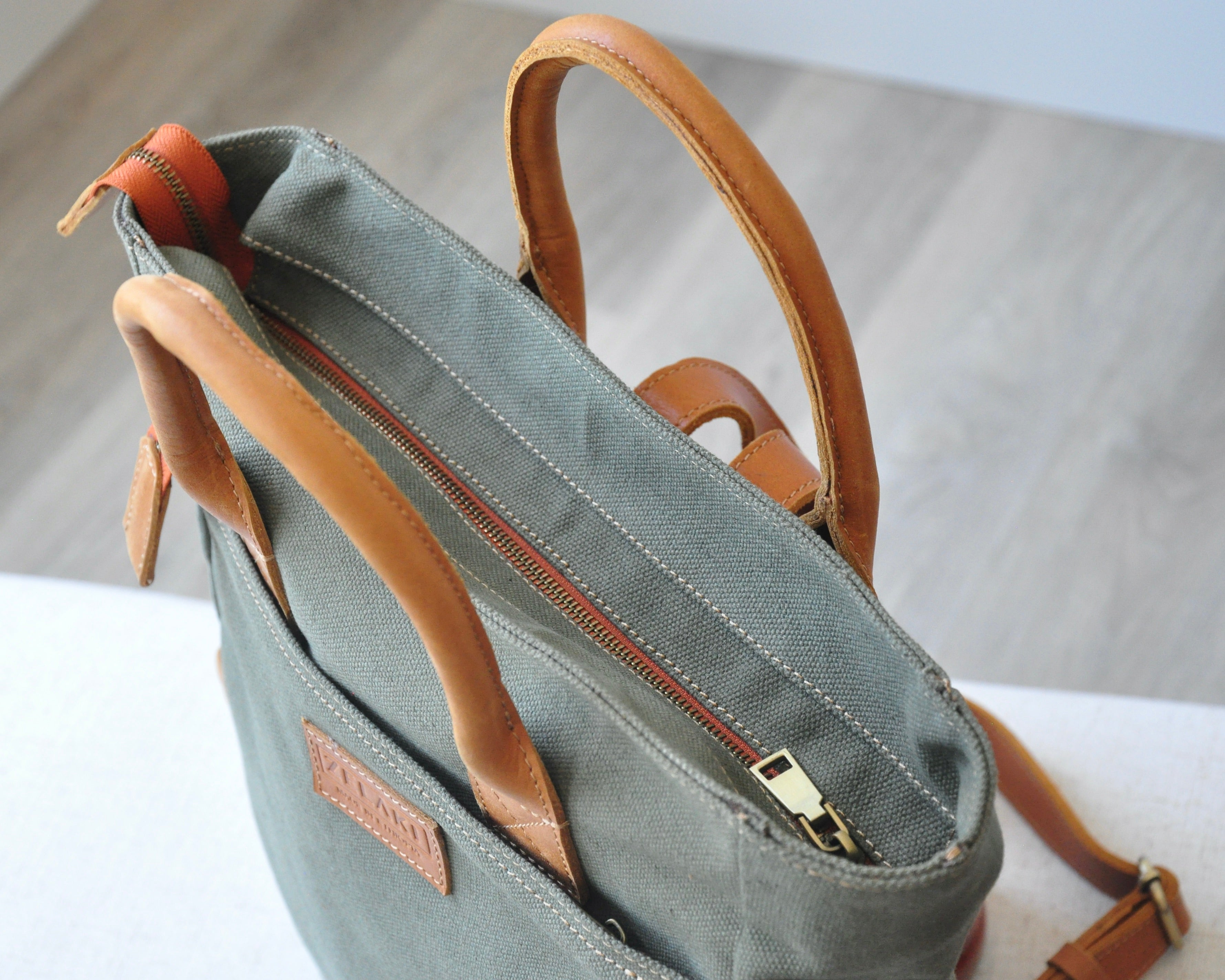 An olive green canvas backpack with full-grain leather accents, featuring two shoulder straps, and top handles.