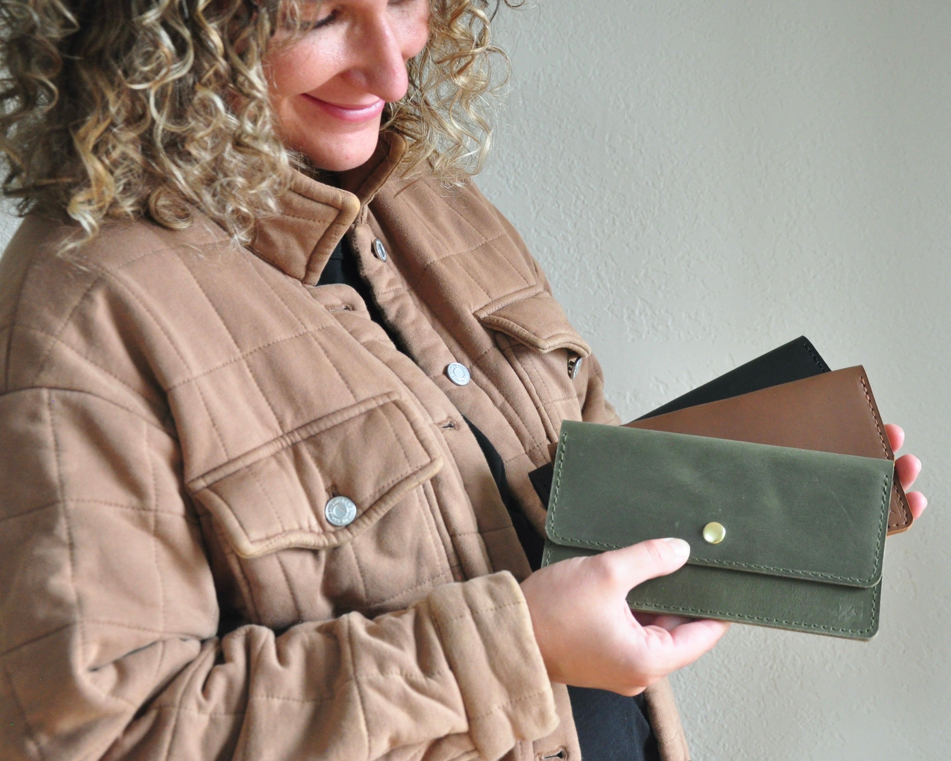 Small business owner holding handmade leather wallet clutches in black, brown, and olive green.
