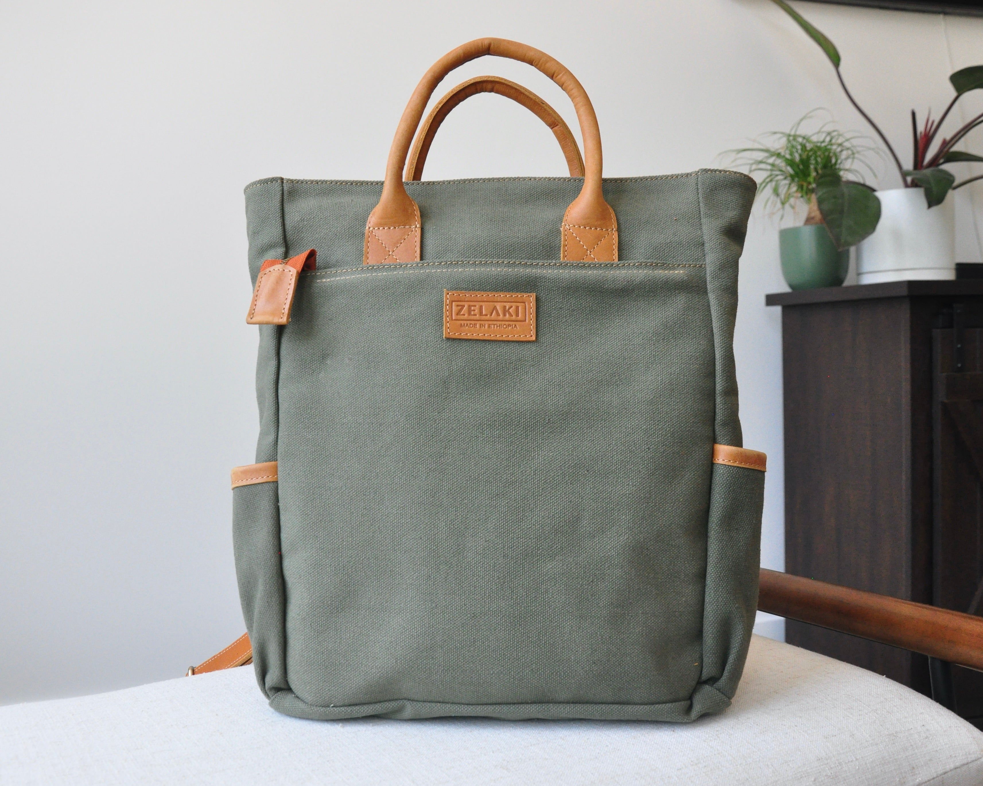 A green canvas backpack with full-grain leather accents, featuring two shoulder straps, a top handle, and placed against a home backdrop.