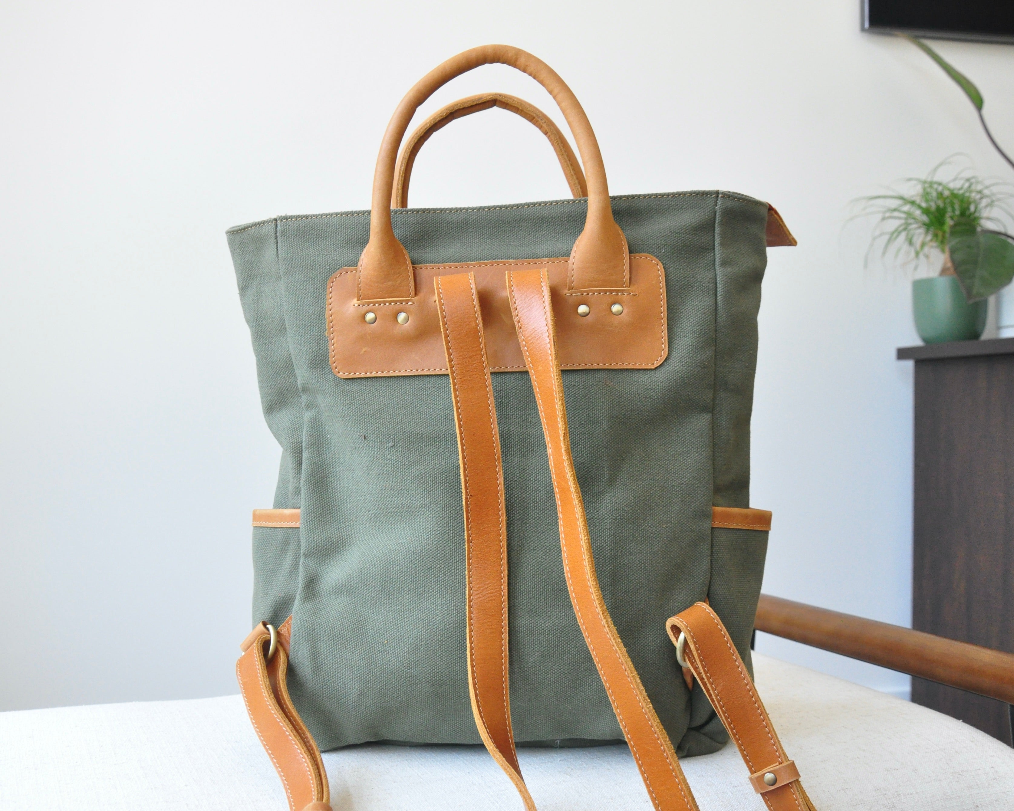 An olive green canvas backpack with full-grain leather accents, featuring two shoulder straps, and top handles.